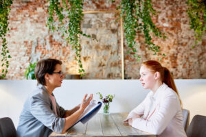 une femme à lunette cadre fait passer un entretien à une jeune femme rousse en face à face. Un mur végétal est présent en fond.
