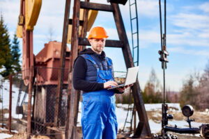 Homme en salopette bleu et avec un casque orange tenant un ordinateur portable dans les mains. Dans le fond une foreuse à pétrole.